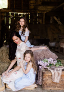 portrait of a mother looking down whilst 2 daughters look to camera in barn by Chelmsford photographer Kika mItchell Photography